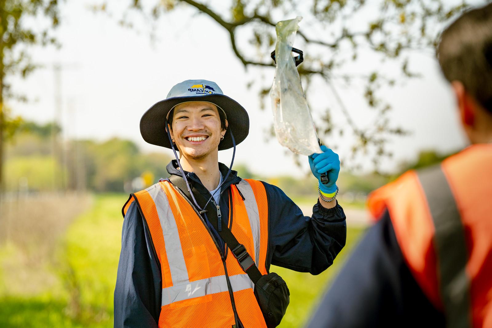 A smiling male holding a plastic bag. He is wearing a safety jacket and a hat. There is someone else in front of him with the same outfit and their outside.